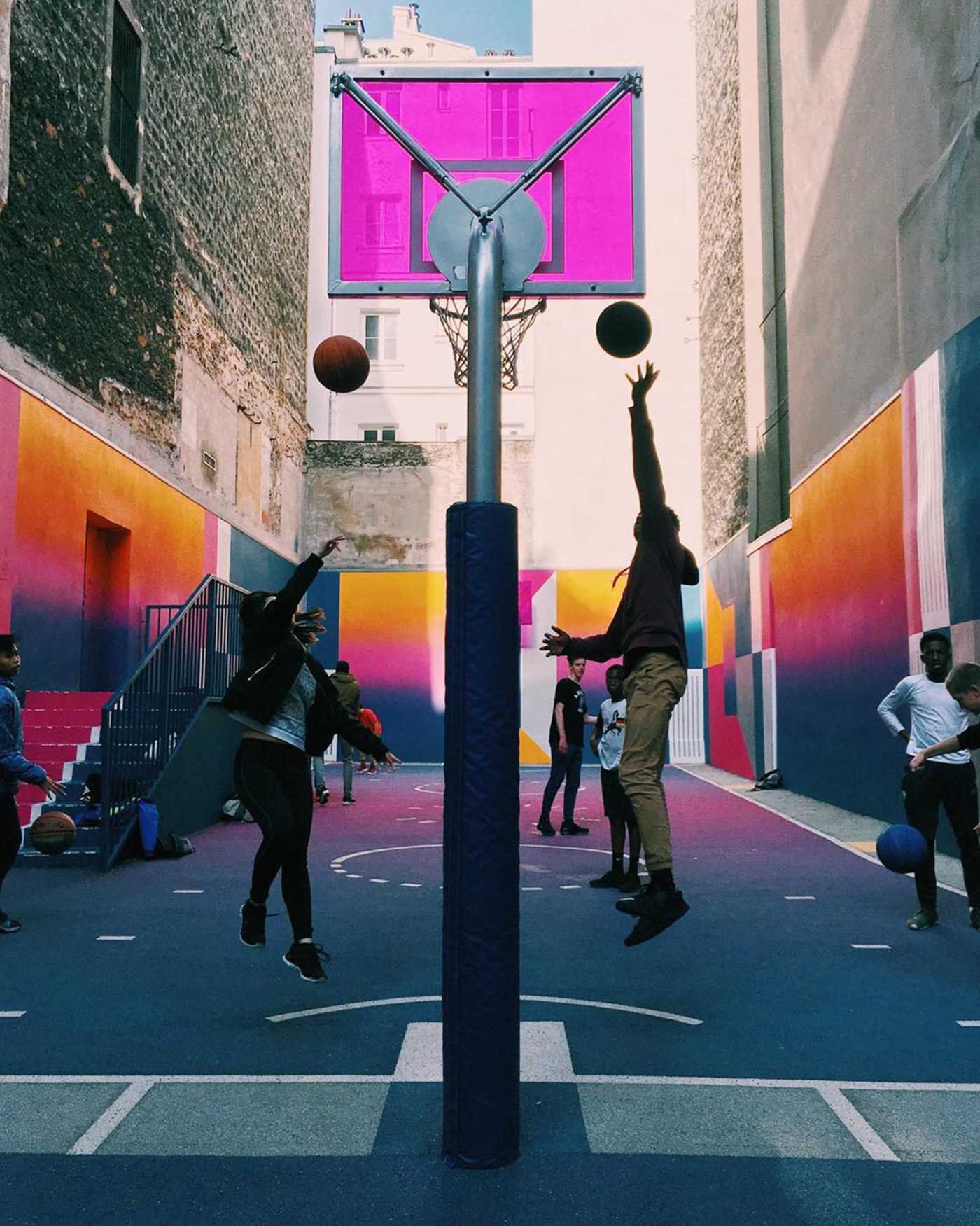 Two people jump to score a basket in an outdoor basketball court against a colorful background